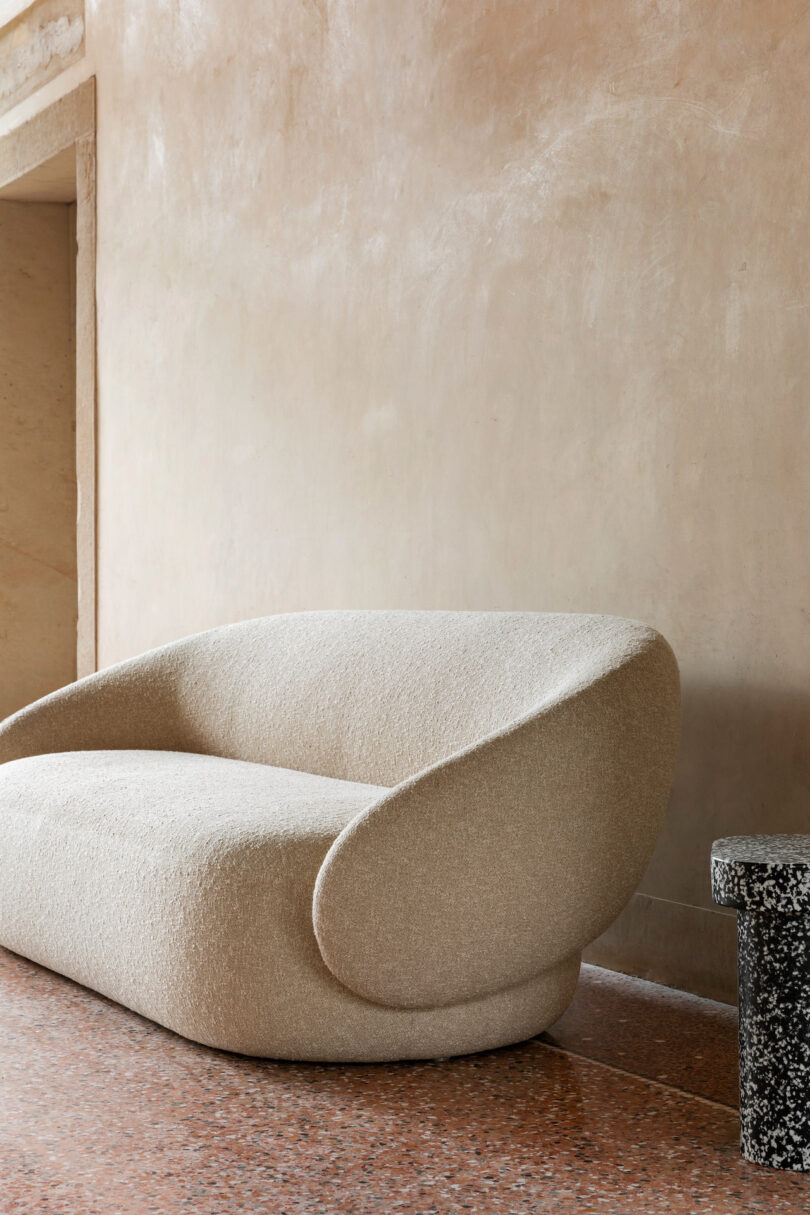 Beige, modern, curved sofa against a textured, neutral-toned wall on a terrazzo floor with a small stone table nearby