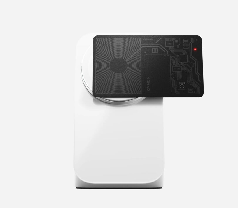 A white wireless charger with a black electronic card on top, featuring circuitry design and a small red light.