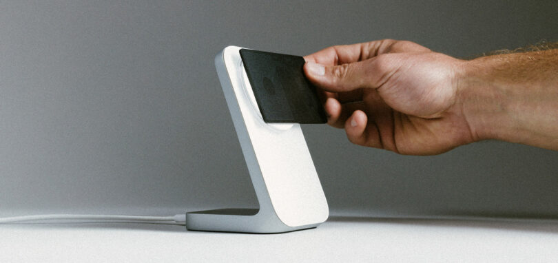 A hand places a small black card on a white, L-shaped charging stand on a gray background.