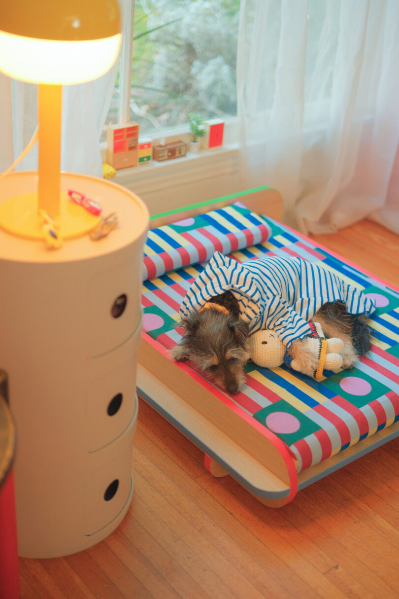 A dog wearing striped pajamas sleeps on a colorful, patterned bed beside a lit lamp in a cozy room