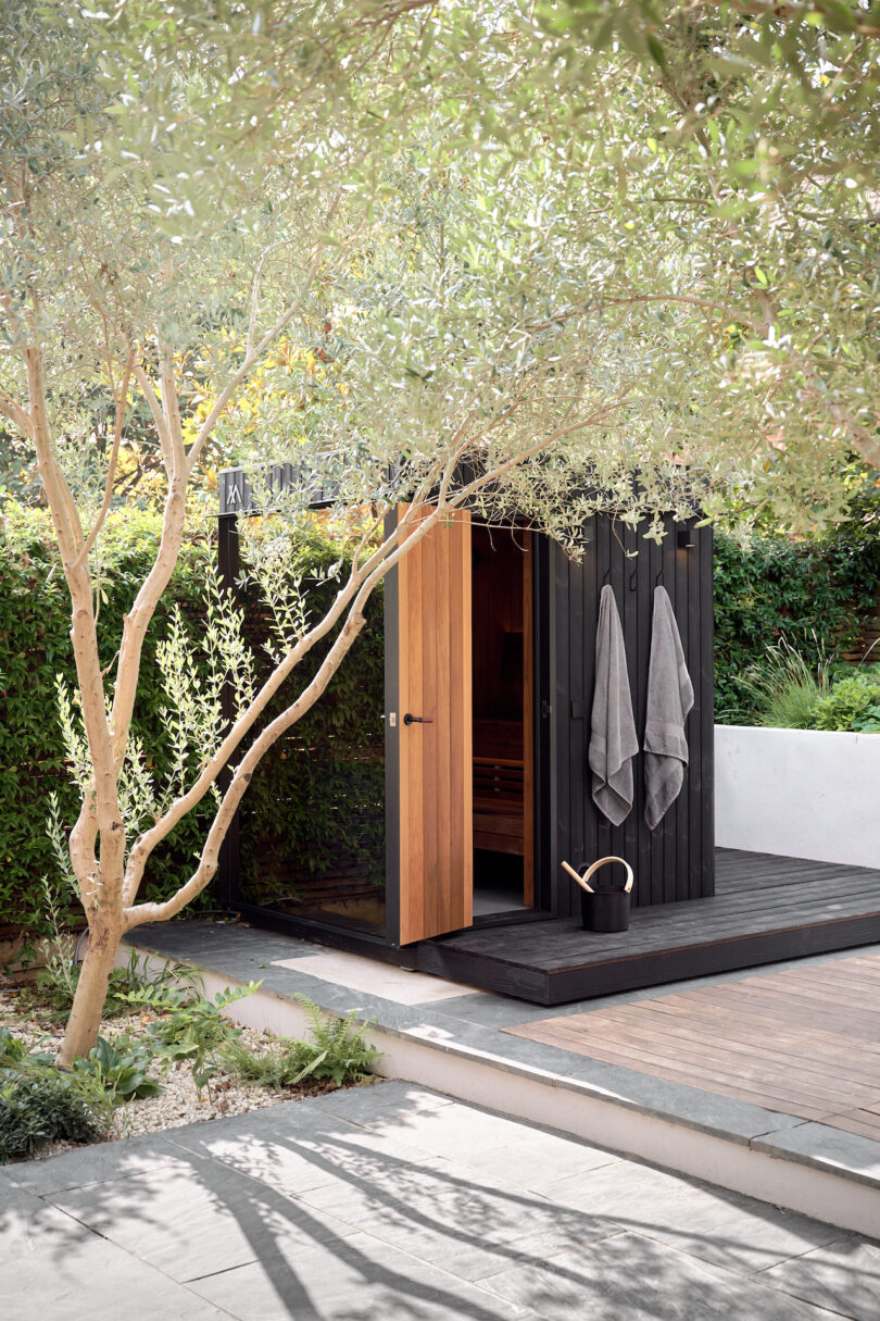 A modern wooden sauna in a garden, with open doors and two towels hanging outside. A watering can is placed nearby, surrounded by trees and greenery