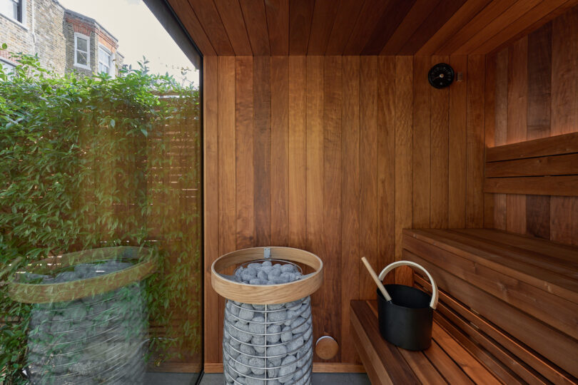 Sauna moderna con paredes y bancos de madera, una cesta de metal con piedras, un cucharón de madera y un cubo negro. Una ventana grande muestra vegetación afuera