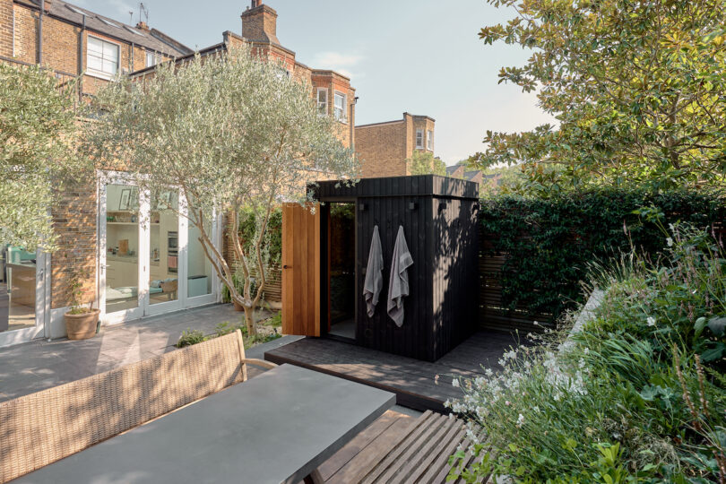 Ducha de jardín al aire libre con recinto de privacidad de madera y toallas colgantes, rodeadas de plantas y árboles, cerca de una casa de ladrillo con grandes ventanas y una mesa de patio