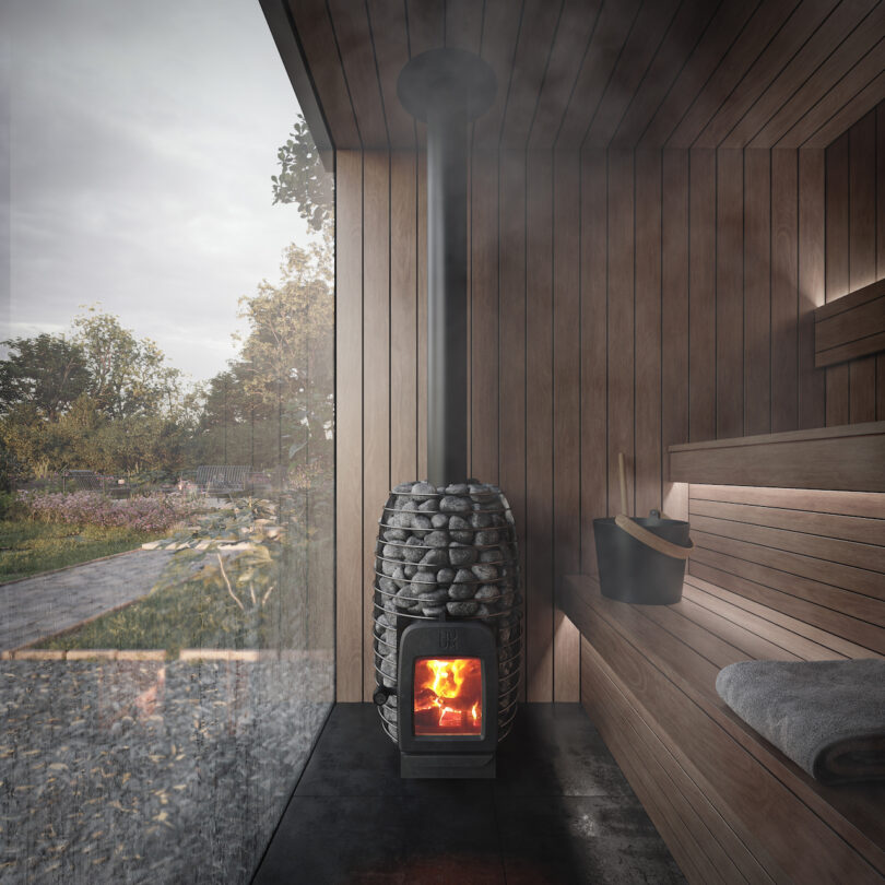 Un interior de sauna de madera con una estufa cubierta de piedra con fuego ardiente. Una toalla y un cubo se sientan en el banco. Una gran ventana de vidrio proporciona una vista del paisaje al aire libre