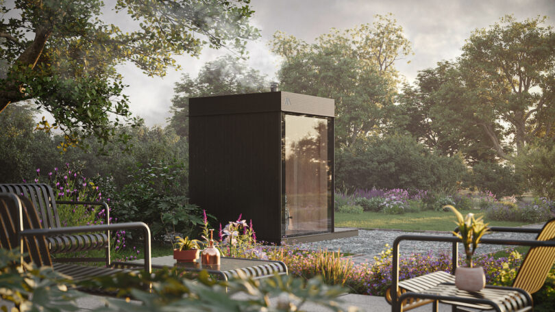 A modern black outdoor sauna is surrounded by lush greenery and flowering plants. Patio furniture with potted plants is in the foreground