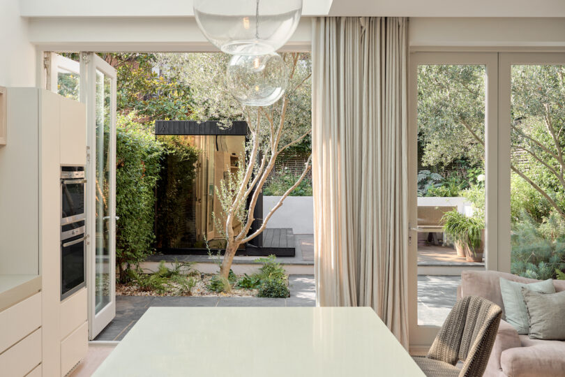 Bright kitchen area with large windows and open doors, leading to a lush garden with trees and plants. Neutral-colored curtains and furniture add to the airy ambiance