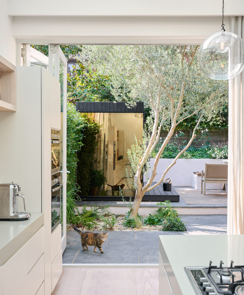 Área de cocina brillante con grandes ventanas y puertas abiertas, lo que lleva a un exuberante jardín con árboles y plantas. Las cortinas y muebles de color neutral se suman al ambiente aireado