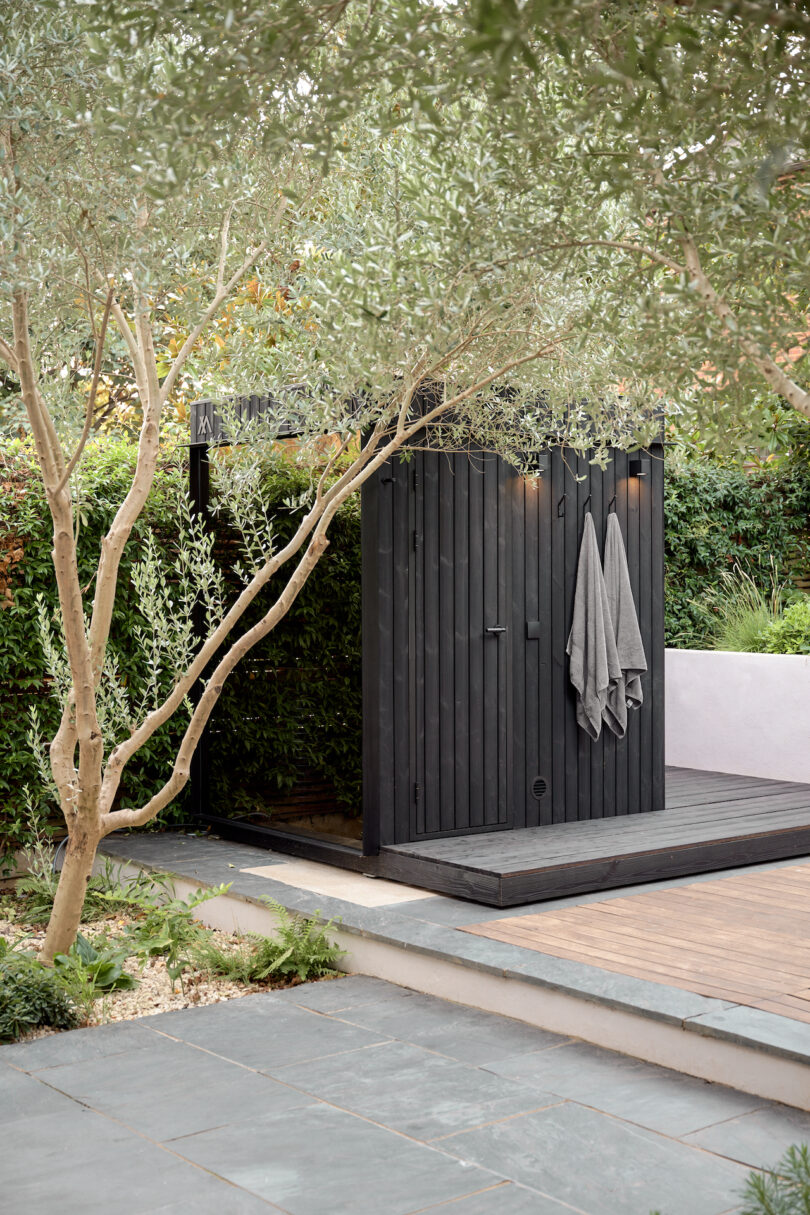 Sauna de madera al aire libre con toallas grises colgadas, rodeadas de vegetación y una pasarela de baldosas en primer plano.