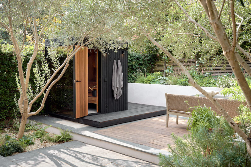 Pequeña sauna de madera con una toalla colgando en la puerta, rodeada de árboles y vegetación, al lado de un banco de mimbre en una cubierta de madera