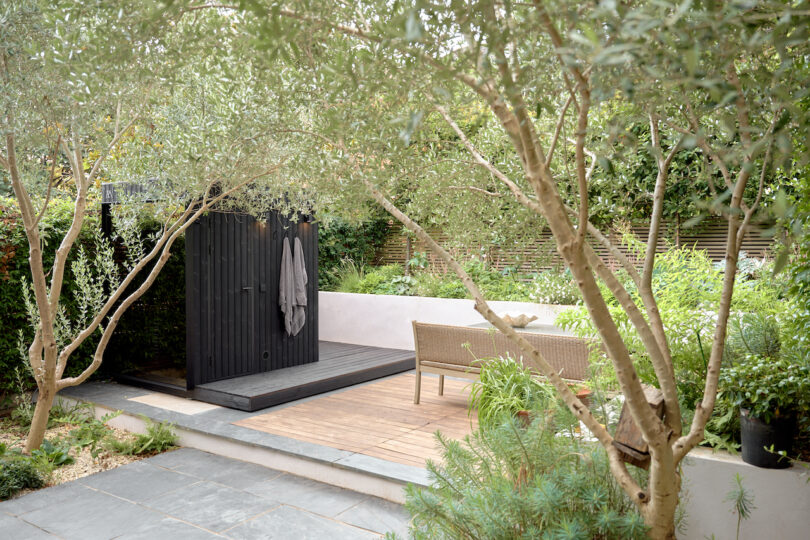 Jardín con árboles y plantas, una terraza de madera, muebles de exterior y una sauna negra al aire libre