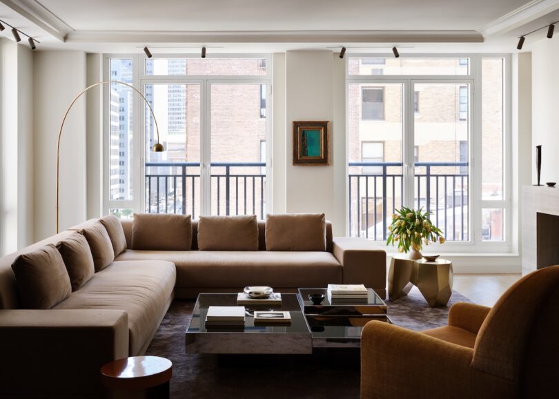 A modern living room with a large beige sectional sofa, a round floor lamp, a glass coffee table, and a potted plant. Large windows let in natural light.