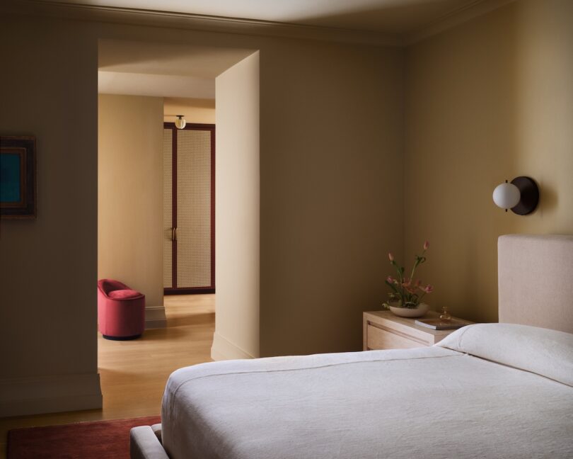 A cozy bedroom with a beige bed and nightstand. A doorway leads to another room with a red chair and closed louvered doors. The walls are painted in neutral tones.