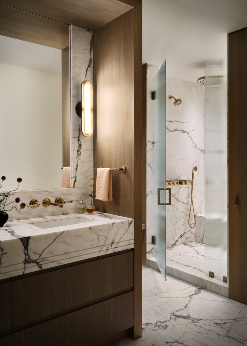 Modern bathroom with a marble sink and shower area. Features wood accents, round wall light, glass shower door, and gold fixtures.