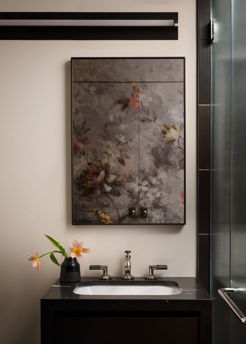 Bathroom vanity with a mirror featuring a floral design. A black sink with a chrome faucet is below the mirror. A black vase with pink flowers is on the left side of the counter.