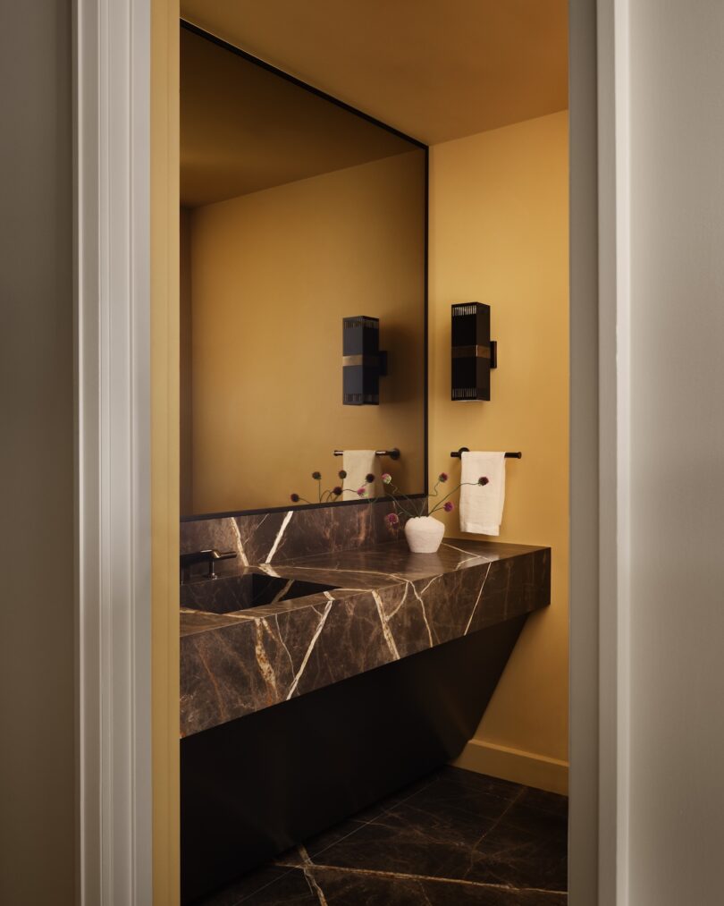 Modern bathroom with a large marble sink, dark tiled floor, and wall-mounted black fixtures. A white vase and hand towel are placed on the countertop.