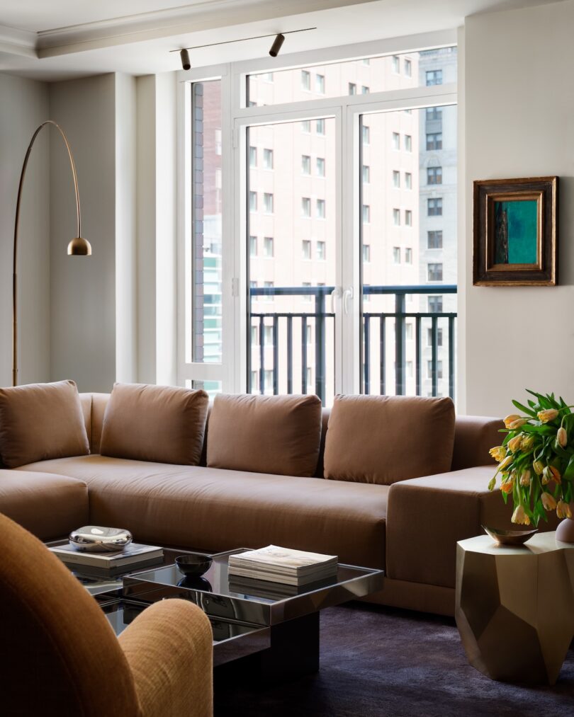 Modern living room with a beige sofa, gold coffee table, and floor lamp. Large window with urban view and a potted plant on the side.