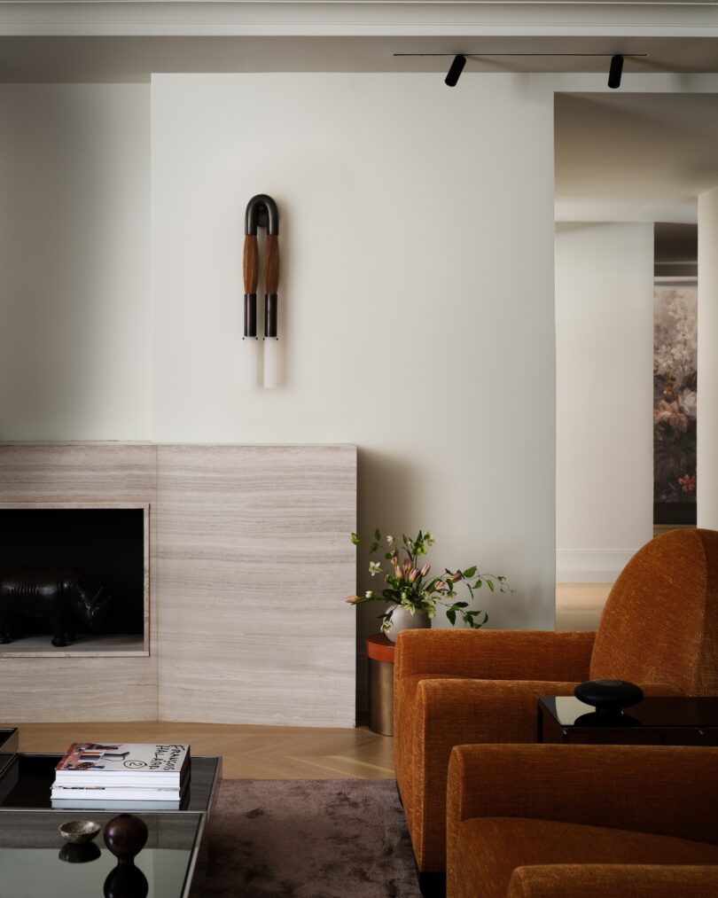 A modern living room with an orange chair, wall sconce, minimalist fireplace, and potted plant. A few magazines rest on a coffee table.