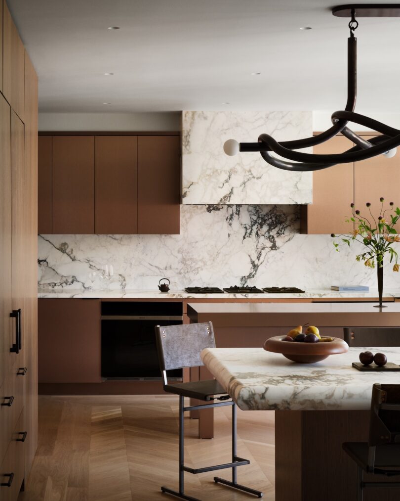 Modern kitchen with brown cabinets, marble countertops, and backsplash. A black chandelier hangs above a marble table with a fruit bowl and a metal chair. A plant is in the corner.