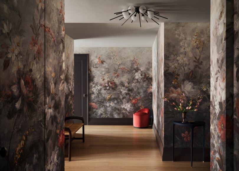 A hallway with floral wallpaper, wooden floor, black side table with a vase, a chair, and a red chair in the corner. Ceiling light fixture with multiple arms is visible.