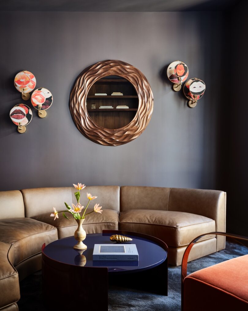 Modern living room with a brown leather sectional sofa, round coffee table, decorative wall sconces, and a round wooden mirror. A vase with flowers sits on the table.