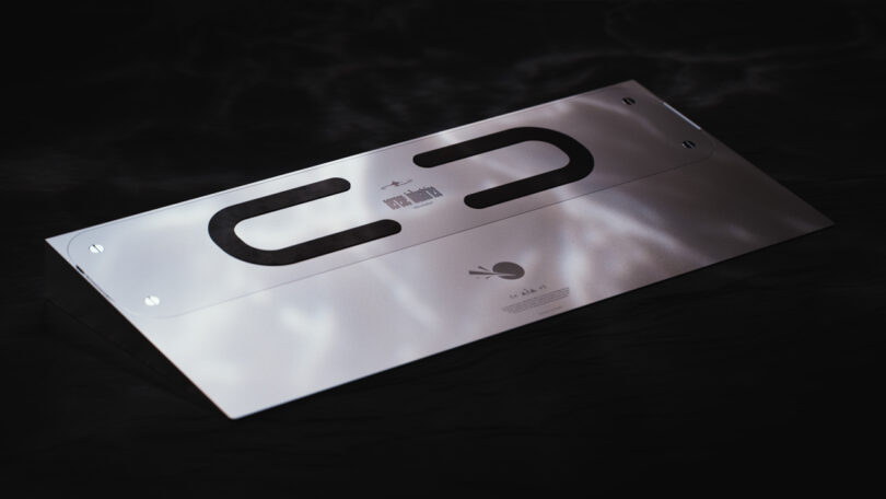 A sleek, metal electronic device with a minimalist design, featuring two long black strips and a small logo on the surface, set against a dark background.