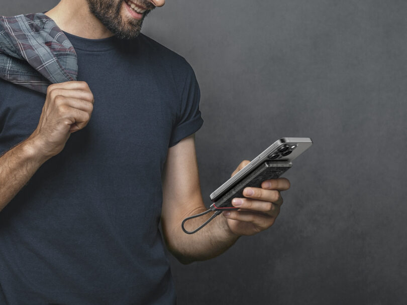 Man in a dark t-shirt holds a smartphone with a strap, slightly smiling and pulling on his jacket over his shoulder against a gray background.