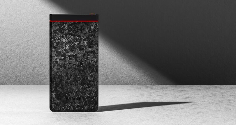 A rectangular, sleek, textured black power bank with a red stripe is on a gray surface, casting a shadow against a light gray wall.