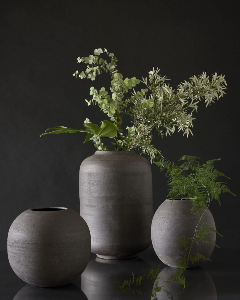 Three gray ceramic vases on a dark background, one large in the center holding greenery, and two smaller empty vases on either side