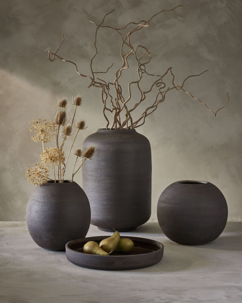 Four dark ceramic vases of varying shapes on a table, with dried flowers and twigs in some. A shallow dish contains two pears