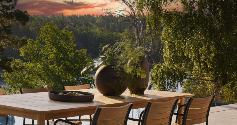 Outdoor dining table with chairs, two large vases with greenery, and a tray with small stones. Surrounded by lush trees with a colorful sunset in the background