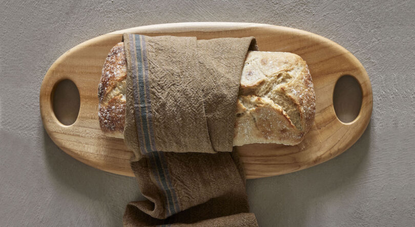 A loaf of bread partially wrapped in a brown cloth rests on a wooden cutting board with handles