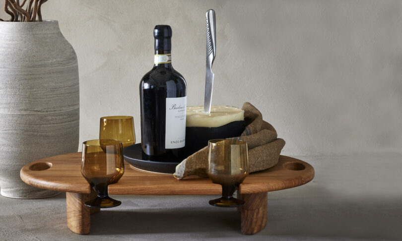 A cheese wheel with a knife, a bottle of red wine, and three amber glasses on a wooden tray