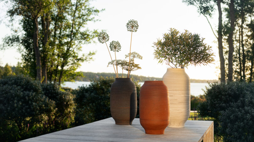 Three ceramic vases with decorative plants on a wooden table outdoors, surrounded by lush greenery and overlooking a distant body of water