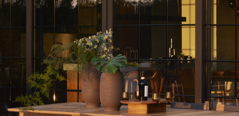 A table with two vases of foliage, a bottle of wine, and glasses is set on a patio with large windows reflecting the evening light