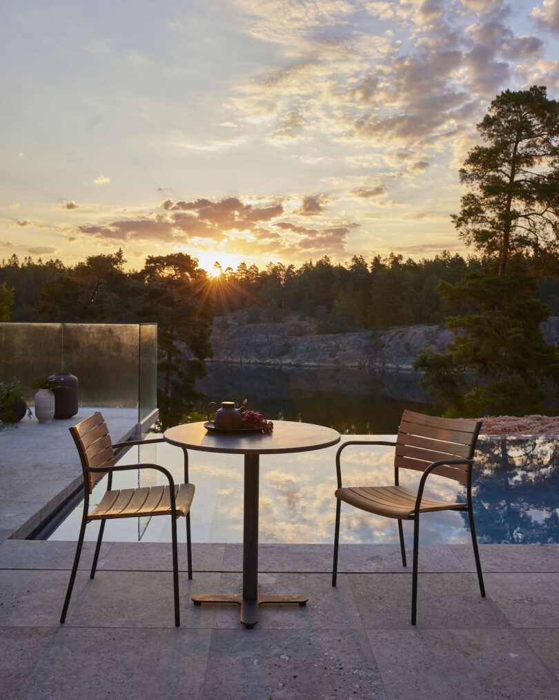 A serene outdoor setting with a table, two chairs, and a teapot by a pool, overlooking a forest and lake at sunset