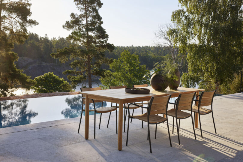 Outdoor patio with wooden dining table, six chairs, and decorative vases. Overlooks a scenic view of trees and water. Sunlit setting with clear skies