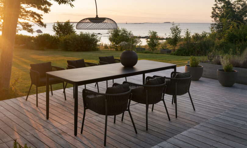 Outdoor dining area on a wooden deck with a black table and chairs, overlooking a serene lake and surrounded by trees and greenery at sunset