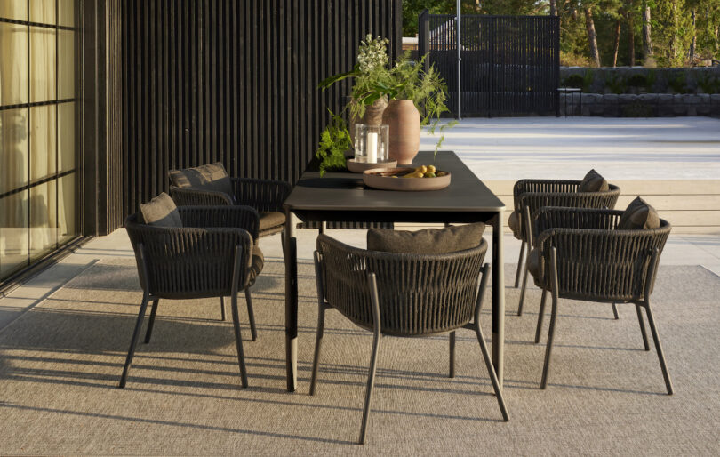 Outdoor dining area with a black table, six black chairs, a large vase, a potted plant, and a tray with items on a light-colored patio