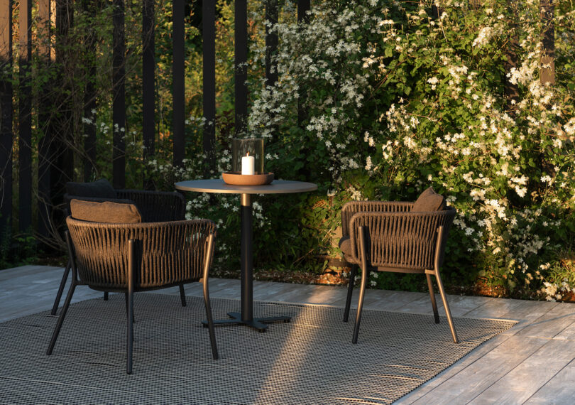 Outdoor patio with two wicker chairs and a round table on a textured rug. A candle sits in a hurricane glass on the table. White flowering shrubs and a slatted fence are in the background