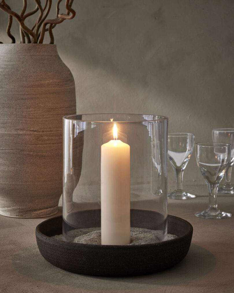 A lit candle in a clear glass holder on a dark tray, next to an earthy vase and empty wine glasses on a table