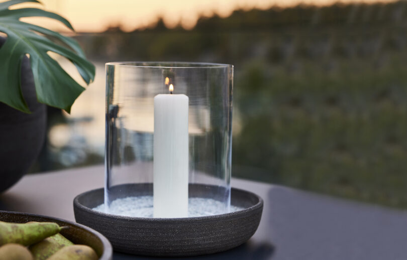 A lit white candle in a glass holder on a table, with a blurred outdoor setting in the background