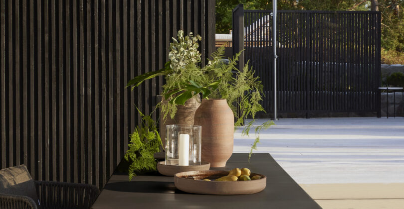 Vases with green foliage, a candle holder, and a bowl of lemons are arranged on a dark table against a black slatted wall