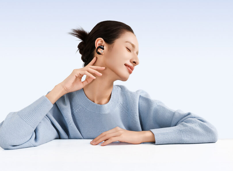 A person wearing wireless earbuds tilts their head and smiles with eyes closed, resting an arm on a white table. They are wearing a light blue sweater.