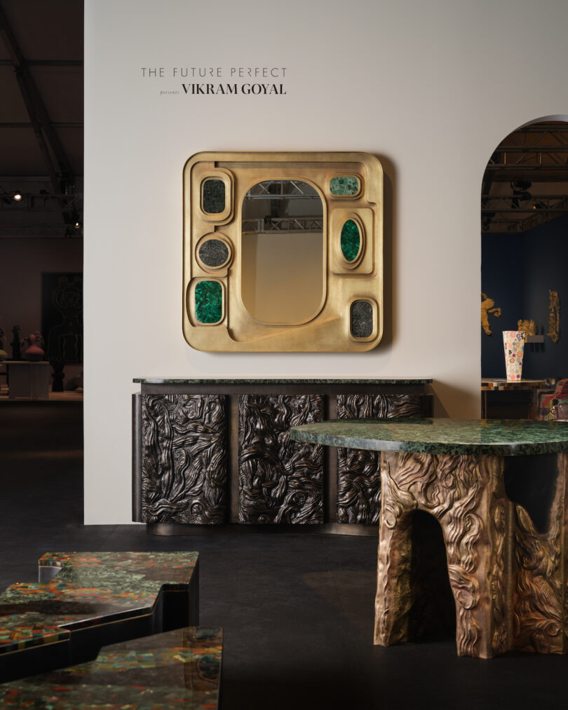 Art exhibit featuring an abstract mirror with gold and green accents, displayed above a textured black cabinet and a table with a sculpted base