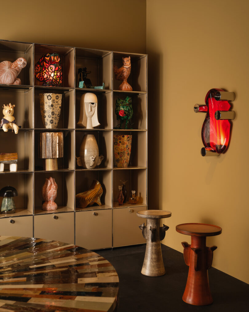 A room with a multi-colored round table, two unique stools, and a shelving unit displaying various artistic objects, including sculptures, vases, and a wall-mounted red guitar art piece