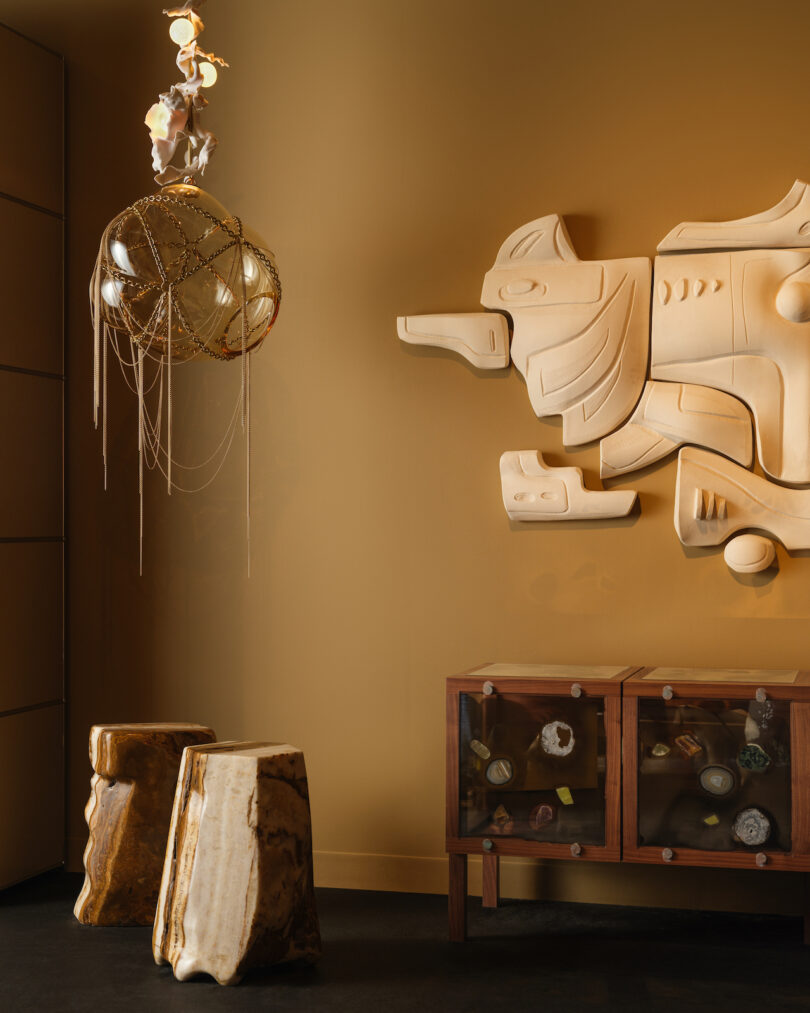 A room features a geometric wall sculpture, a spherical pendant light, and a wooden cabinet with glass doors. Two stone-like stools are in the foreground, all set against a warm mustard wall