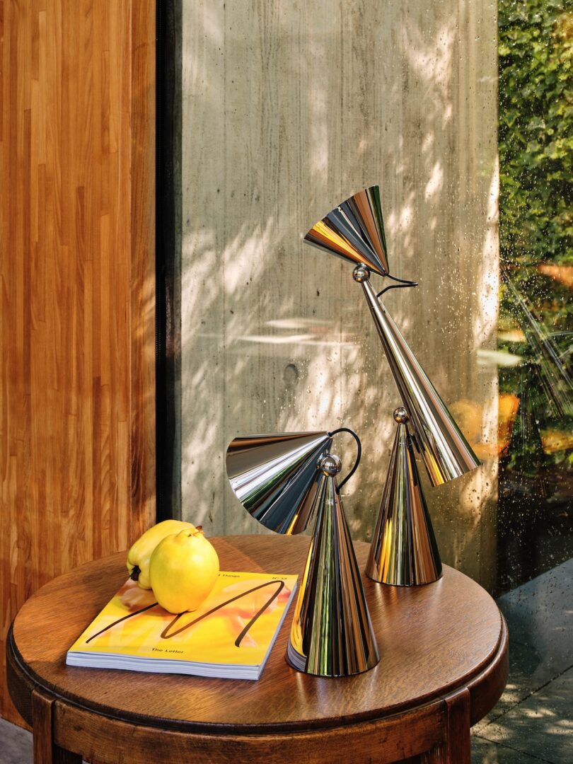 Modern metallic sculptures and a yellow fruit on a wooden table, next to a bright and open window with a leafy view outside