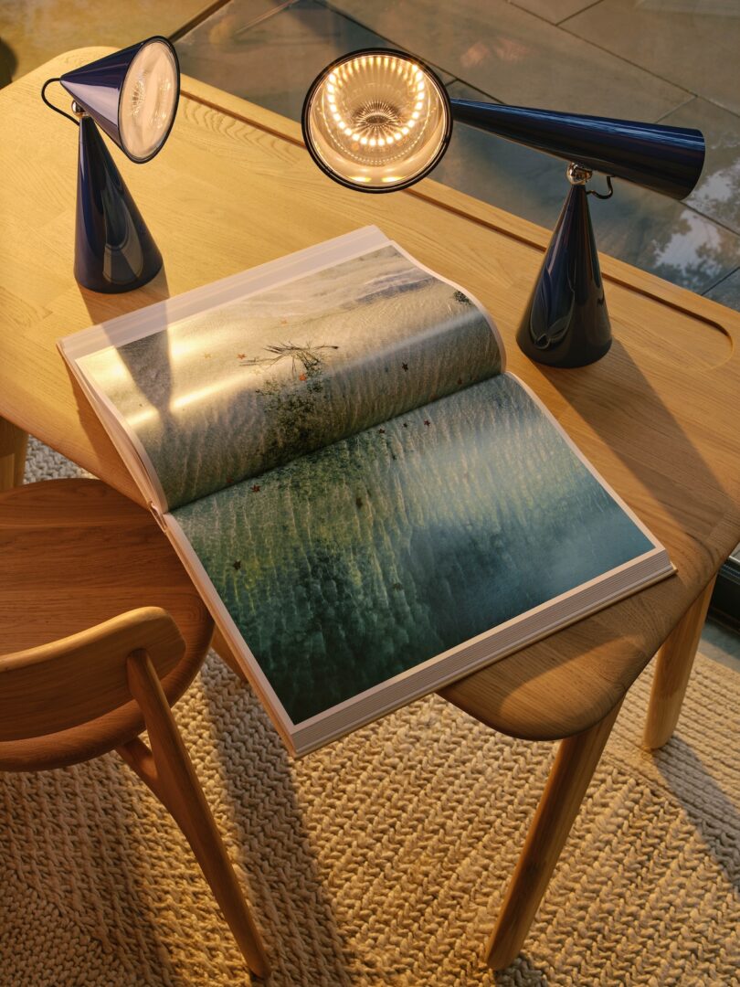 A wooden table with an open book displaying a beach scene, two modern black lamps, and a wooden chair