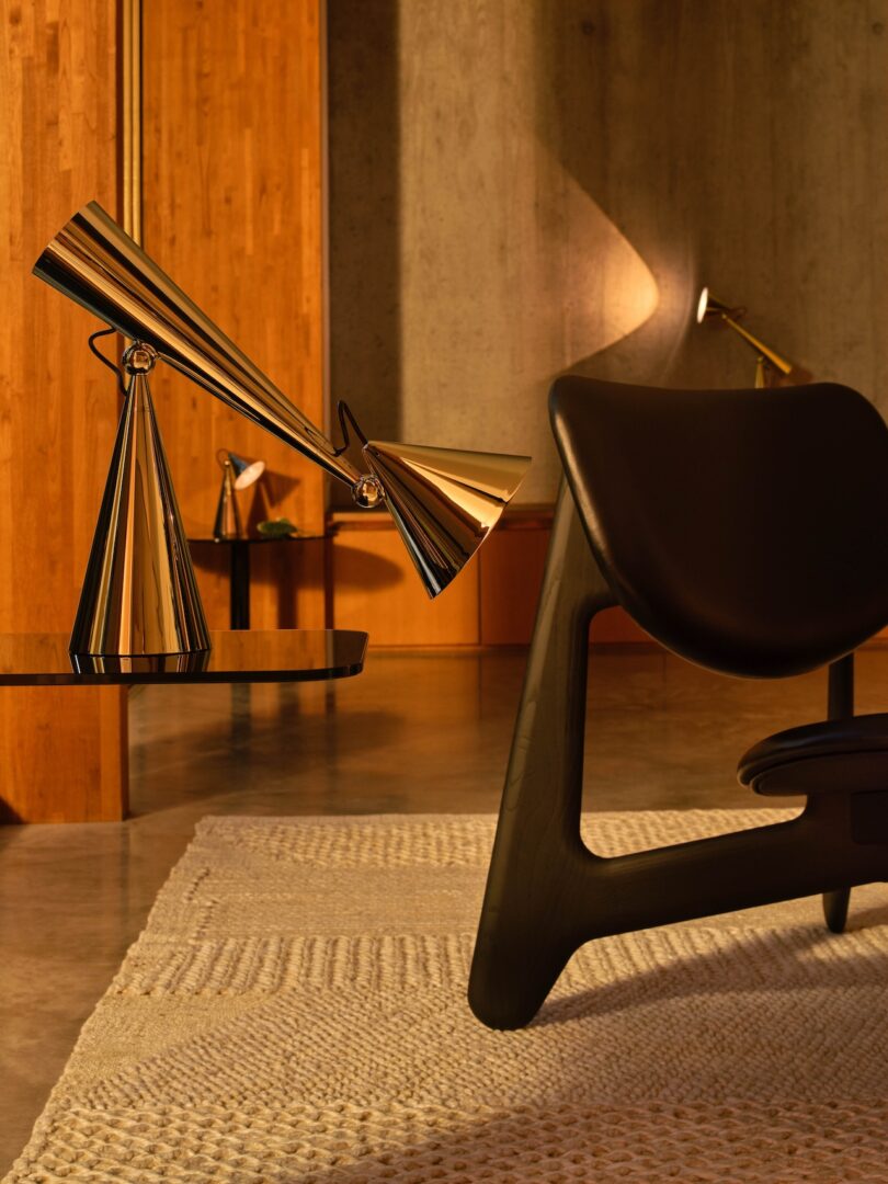 A modern room with a black chair, a textured rug, and two geometric gold lamps on a glass table. Warm lighting enhances the wooden wall and concrete backdrop