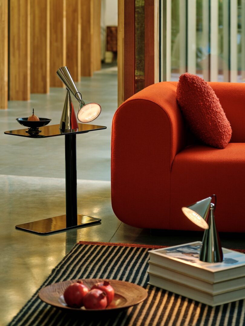 A modern living room with a red couch, a black side table with a silver lamp, and a striped rug. Apples are in a bowl on the rug, and books are stacked on the table